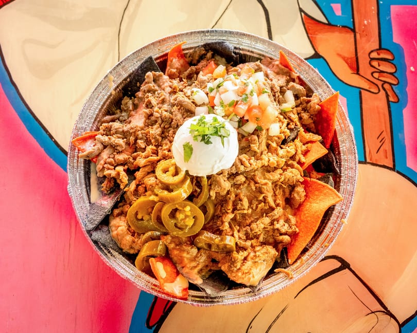 Loaded nachos with ground meat, pico de gallo, sour cream, and jalapeños in a foil container.