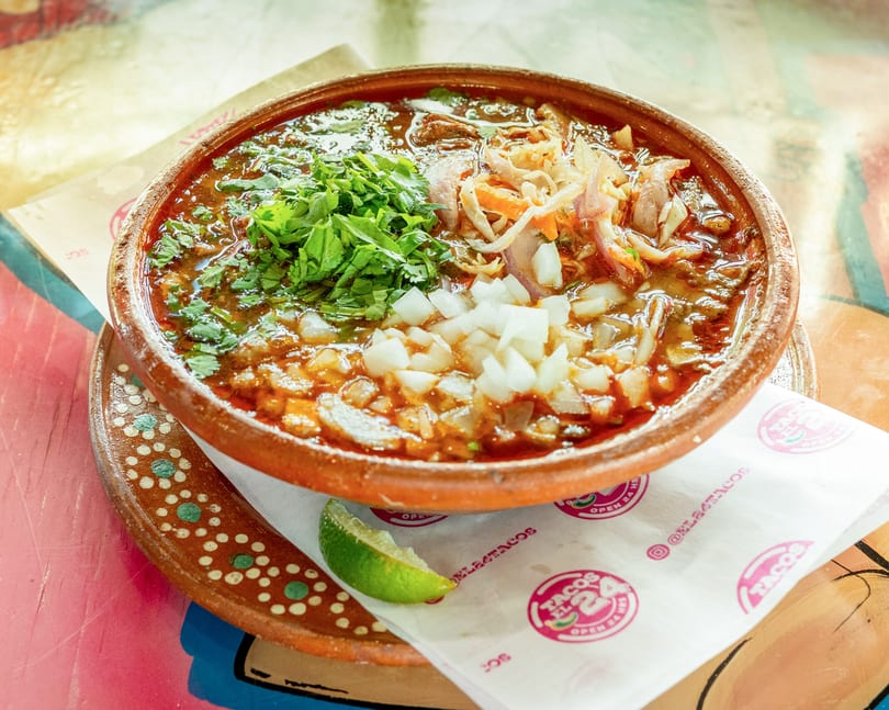 A bowl of Tacos El 24 birria stew with cilantro, onions, and a lime wedge.