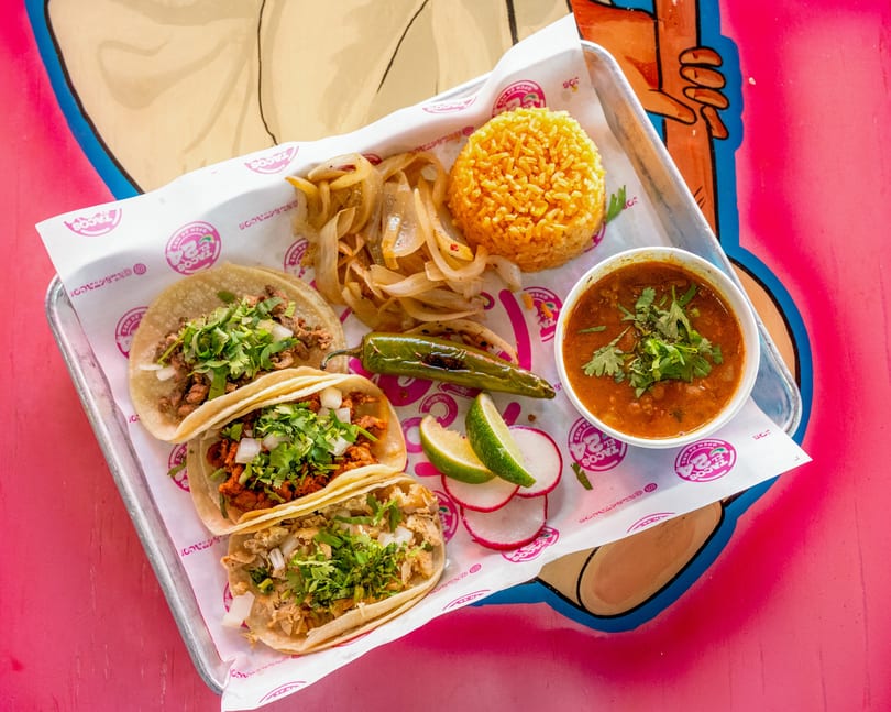 Three tacos, rice, grilled onions, a chile, limes, radishes, and a bowl of soup on a tray.