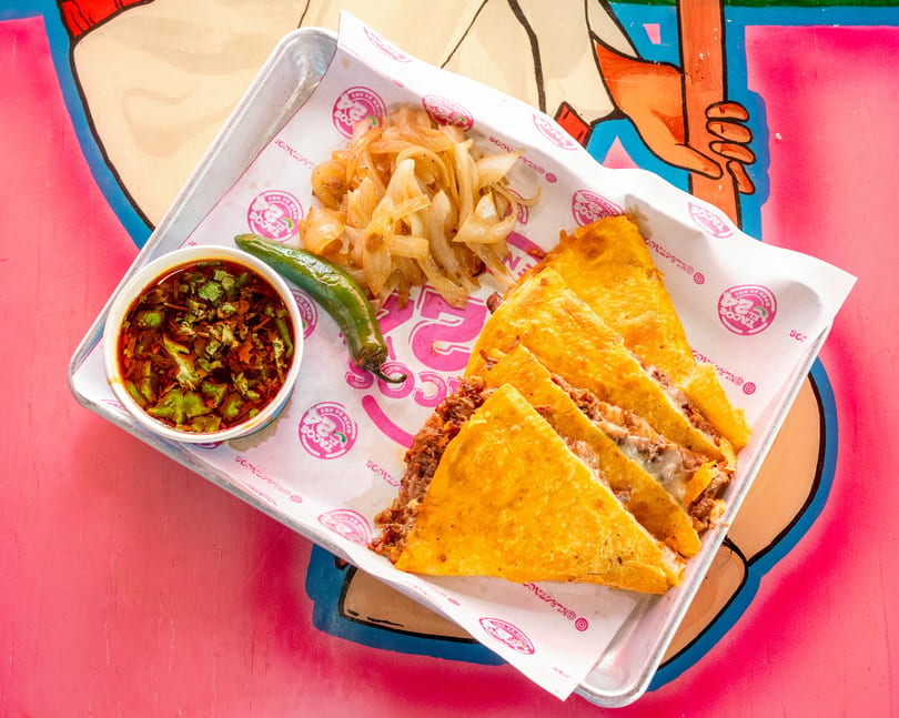 Birria quesadillas with consome, grilled onions, and a serrano pepper on a metal tray.