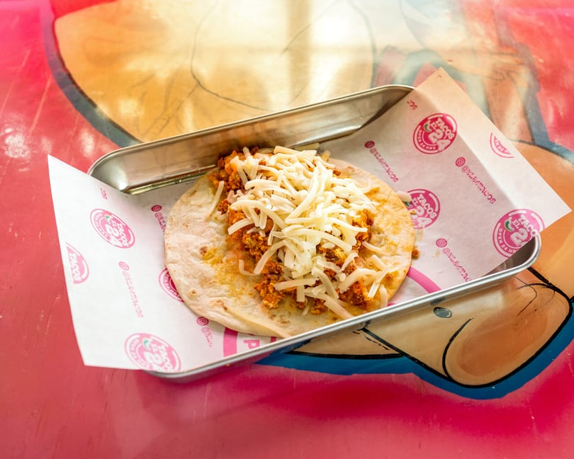 A Tacos El 24 taco with chorizo and shredded cheese on a tortilla, served in a metal tray.
