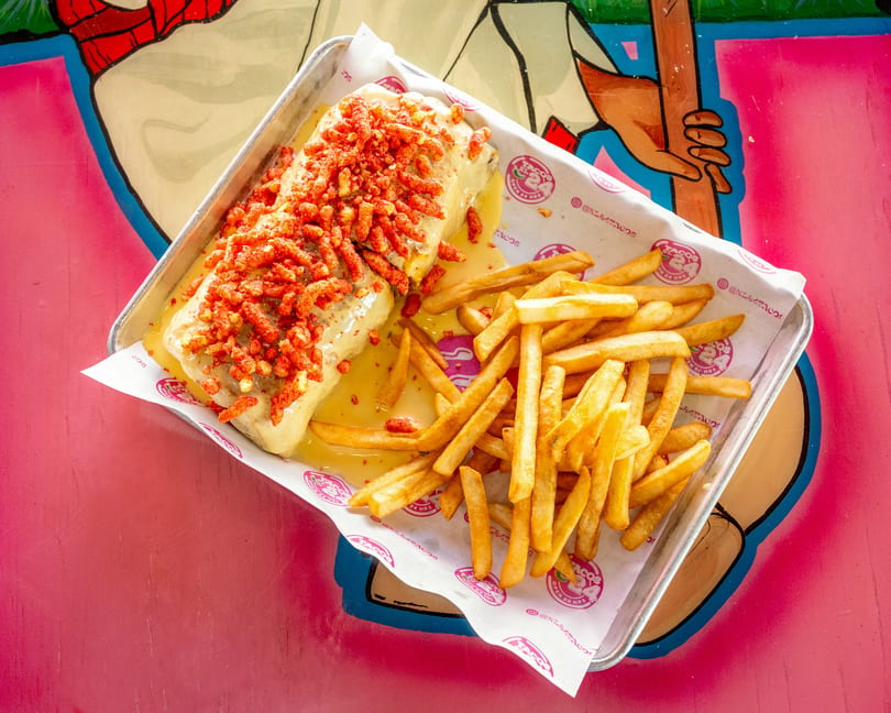 Burrito covered in cheese sauce and red crunchy toppings, served with golden fries on a metal tray.